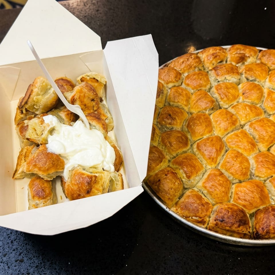 Golden-brown baked pastries fill a round pan beside a takeout box of pastry pieces topped with whipped cream.